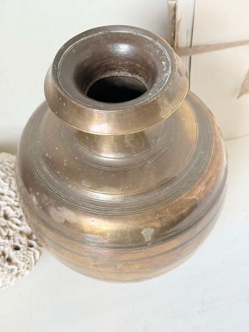 An antique brass-colored metal vase with a rounded body and a flared rim, shown on a light surface with a lace doily and neutral background.