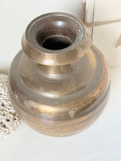 An antique brass-colored metal vase with a rounded body and a flared rim, shown on a light surface with a lace doily and neutral background.