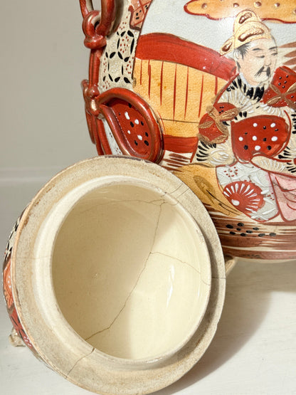 A close-up of a cracked ceramic jar with its lid leaning against the body. The jar has a hand-painted traditional illustration featuring a figure in red, orange, black, and white, with a cream-colored interior visible on the open lid.