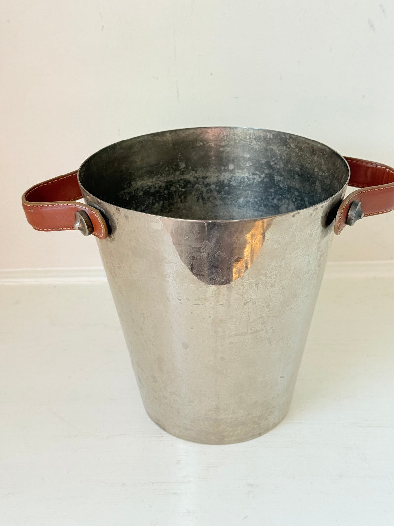 Vintage metal wine cooler with leather handles and tapered form, photographed on white surface.