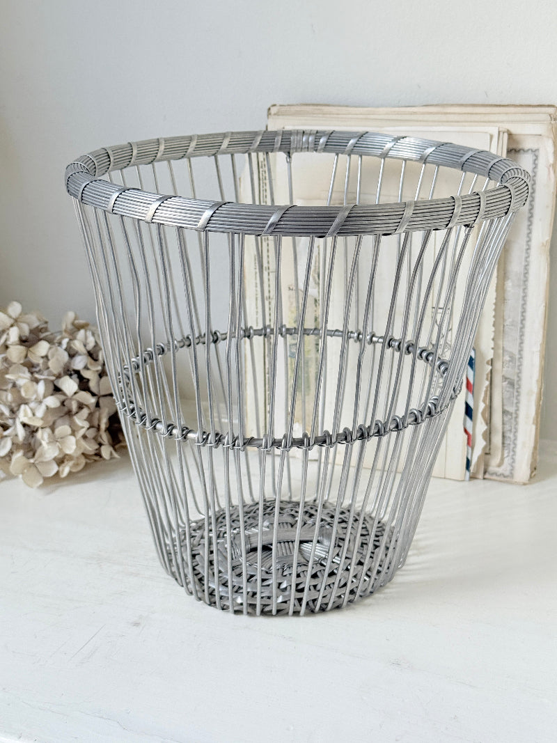 A silver metal wire basket with a woven circular design, displayed on a white surface with vintage-style frames and dried flowers in the background.