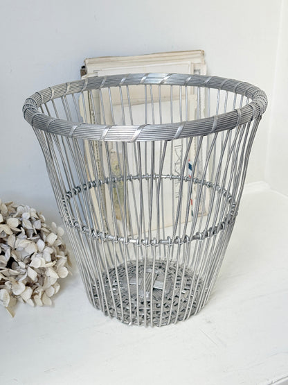 A silver metal wire basket with a woven pattern and a thick top rim, styled on a white surface with dried flowers and a book in the background.