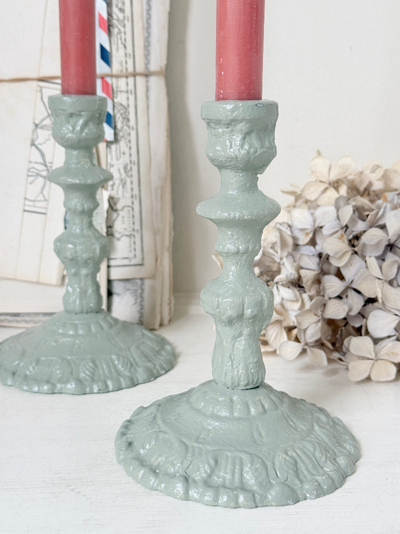 Two ornate cast iron candlesticks in a muted sage green finish, holding pink candles, styled on a white surface with vintage maps and dried flowers in the background.