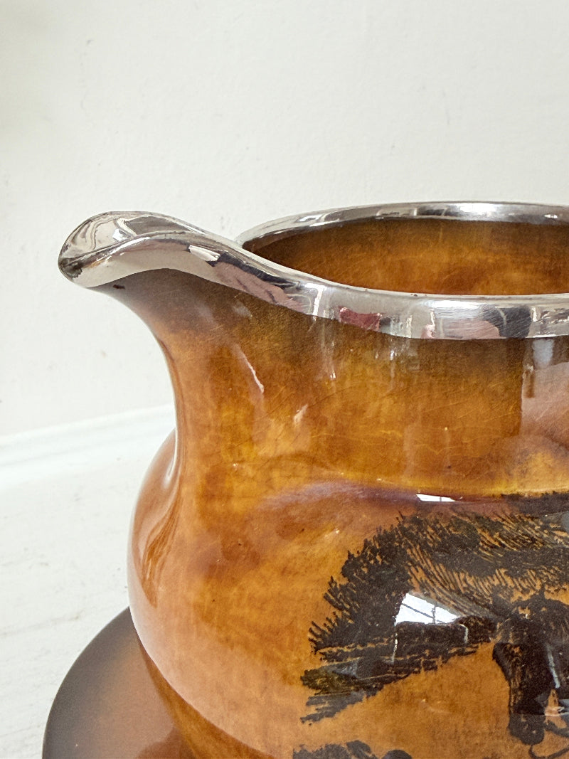 Antique Ridgways stoneware creamer jug and saucer photographed on a white surface in amber treacle glaze with black coaching scenes and silver lustre rim
