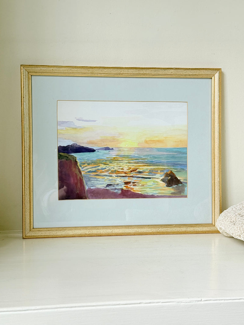 Framed coastal watercolour of sunset over sea with rocks in foreground, in gilt frame with pale mount, photographed on white surface.