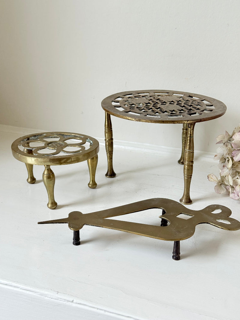 Vintage brass trivet trio with ornate cutwork designs and aged patina, photographed on a white surface