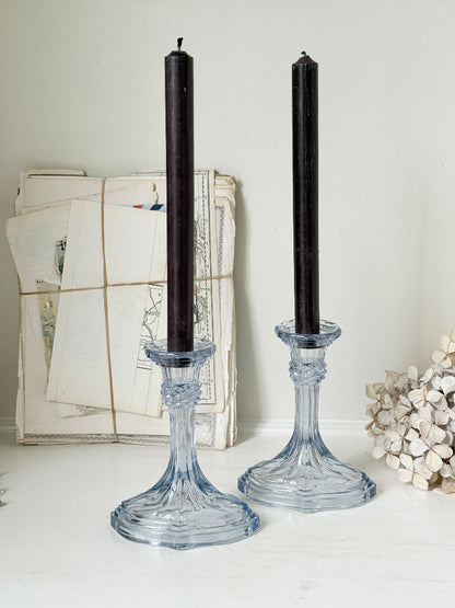 Two tall black taper candles in clear glass candlestick holders, displayed on a white surface against a light wall with vintage papers and dried flowers in the background.