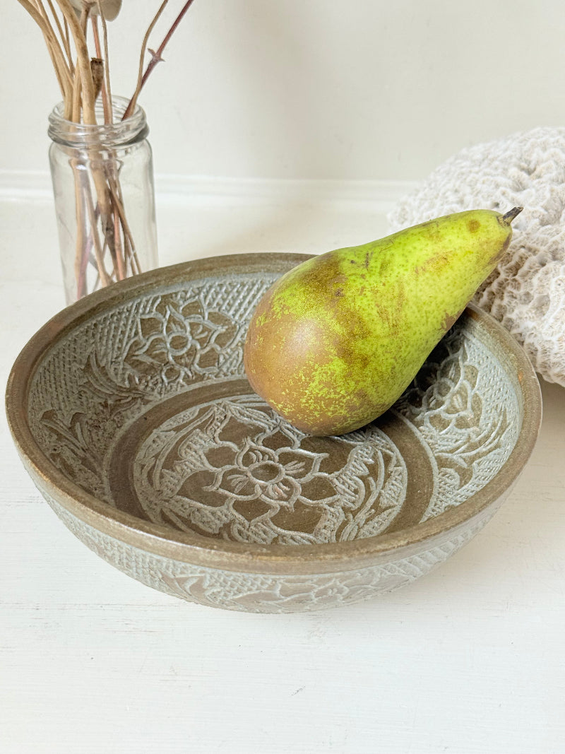 Studio pottery bowl with carved floral and geometric decoration in neutral glaze, photographed on a white surface with a pear inside.