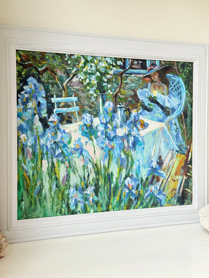 Framed impressionist-style oil painting of blue irises and garden scene, photographed on a white surface