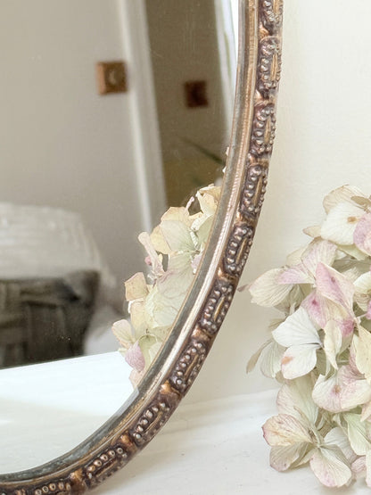 Vintage oval wall mirror with decorative gilt frame and bow crest, styled against a neutral wall with hydrangeas.