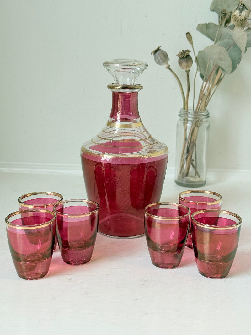 Vintage red printed glass cocktail set with clear glass decanter and six small glasses with gilt band rims, photographed on a white surface