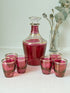 Vintage red printed glass cocktail set with clear glass decanter and six small glasses with gilt band rims, photographed on a white surface