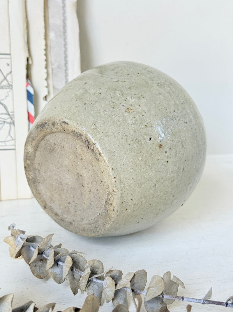 A handmade ceramic planter pot with a textured, speckled light gray-green glaze, shown lying on its side on a white surface with dried eucalyptus leaves in the foreground.