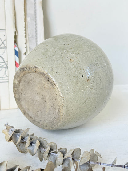 A handmade ceramic planter pot with a textured, speckled light gray-green glaze, shown lying on its side on a white surface with dried eucalyptus leaves in the foreground.