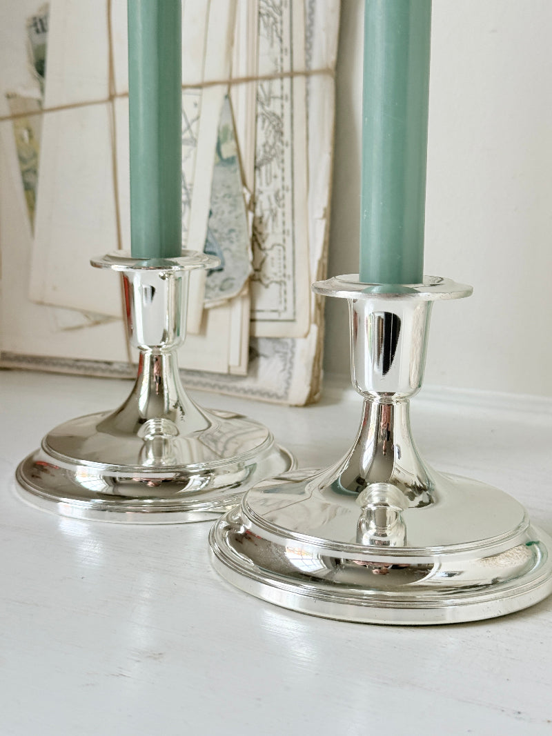Pair of silver tone candlesticks with wide round stepped bases holding green candles, photographed on a white surface