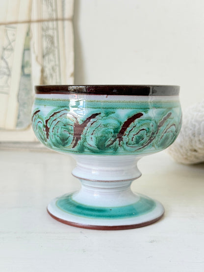 Hand thrown studio pottery footed bowl with green and brown swirling decoration photographed on a white surface