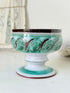 Hand thrown studio pottery footed bowl with green and brown swirling decoration photographed on a white surface