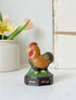 A small hand-painted decorative rooster figurine on a dark base with a green grassy platform, displayed on a white surface with a white vase of tulips and a book in the background.