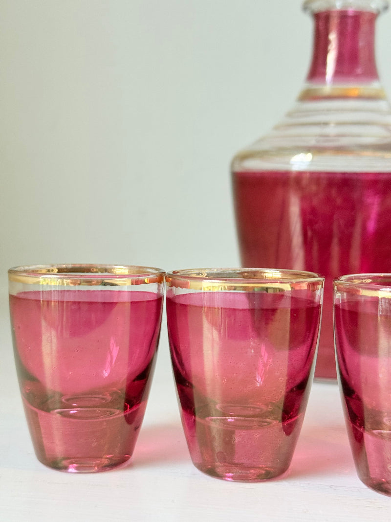 Vintage red printed glass cocktail set with clear glass decanter and six small glasses with gilt band rims, photographed on a white surface