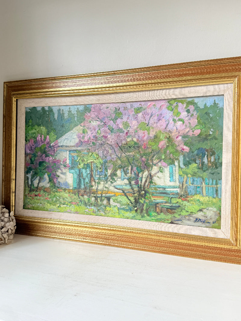 A framed oil painting of a garden scene with pink flowering trees, a white house, and green foliage, housed in a wide ornate gold frame and displayed on a white surface against a white wall.