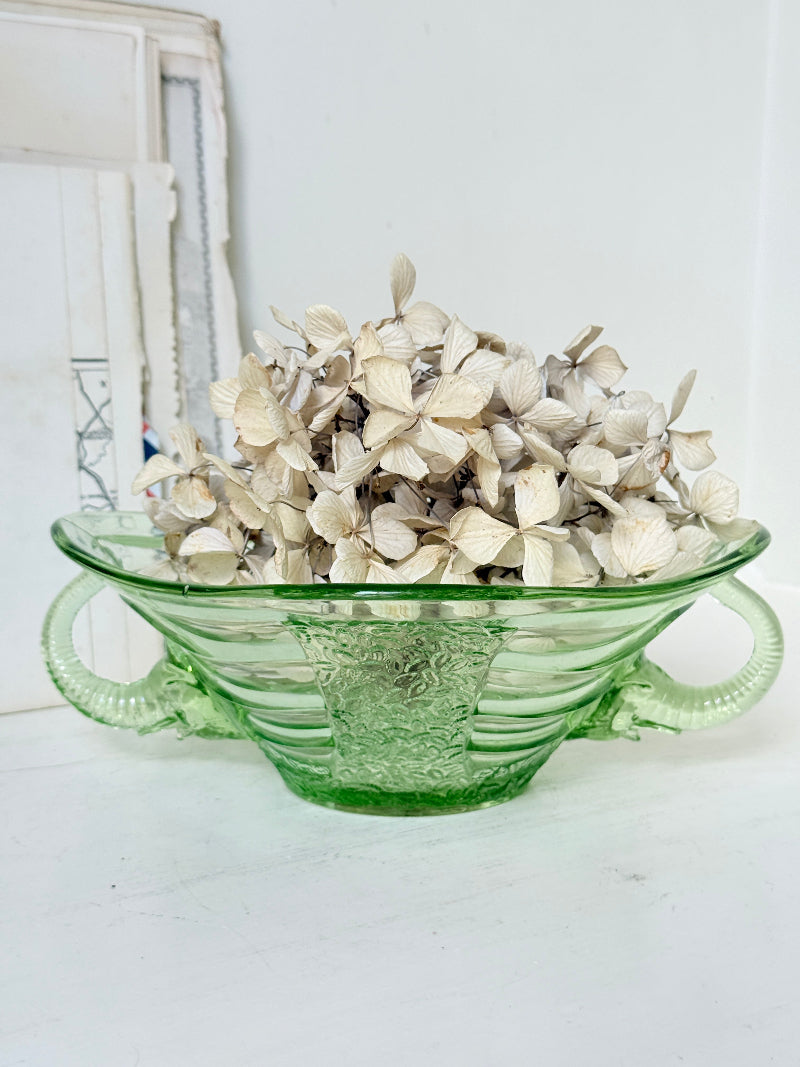 A green glass bowl with side handles, filled with dried cream-colored flowers, set against a white background with a rustic white frame visible on the left.