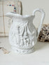 A white ceramic pitcher with intricate embossed classical figures and a draped scene, set against a softly styled background with vintage mail and dried flowers.