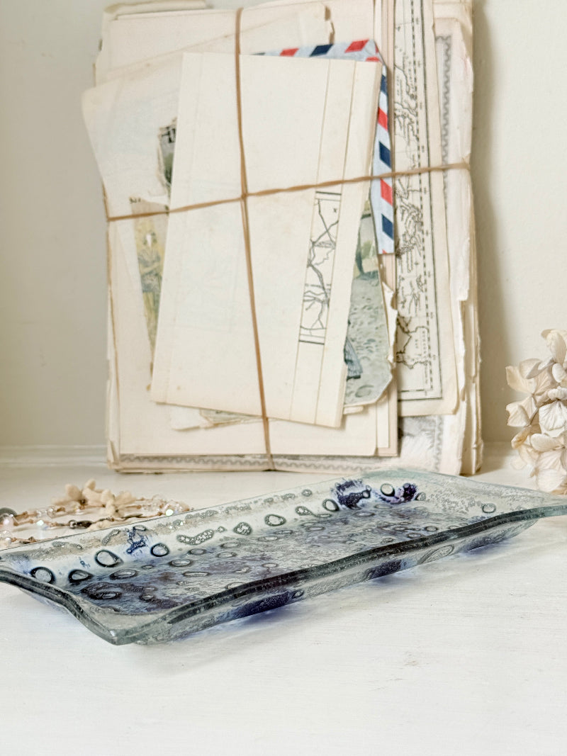 A rectangular glass tray with a wavy edge, featuring a mottled blue and gray pattern with a raised circular border, displayed on a white surface in front of a stack of vintage letters tied with string.