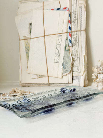 A rectangular glass tray with a wavy edge, featuring a mottled blue and gray pattern with a raised circular border, displayed on a white surface in front of a stack of vintage letters tied with string.
