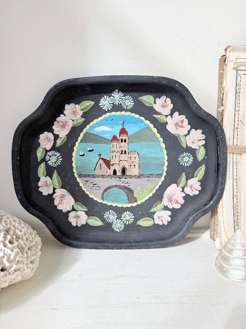 A black decorative tray with scalloped edges, featuring a hand-painted illustration of a castle in a lake surrounded by pink flowers and white daisies, displayed on a white surface.