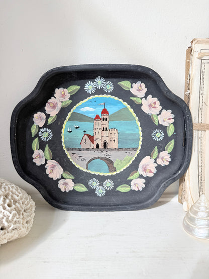 A black decorative tray with scalloped edges, featuring a hand-painted illustration of a castle in a lake surrounded by pink flowers and white daisies, displayed on a white surface.