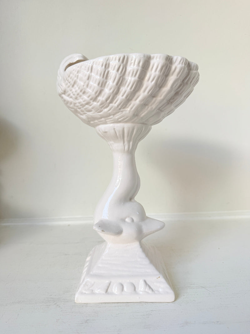 A white ceramic decorative pedestal bowl with a sculpted neck and base, photographed against a plain off-white wall and surface.