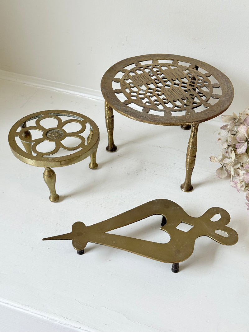 Vintage brass trivet trio with ornate cutwork designs and aged patina, photographed on a white surface