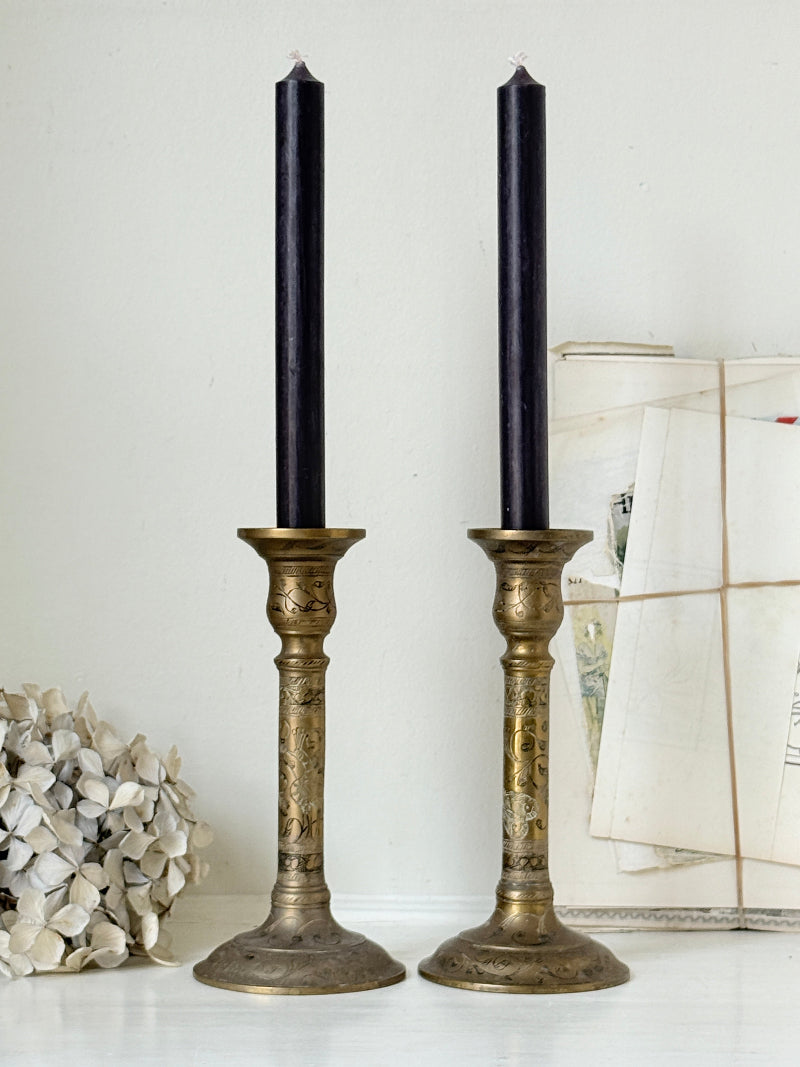 Pair of tall Indian engraved brass candlesticks in the Benares style with floral detailing and black taper candles on a white surface