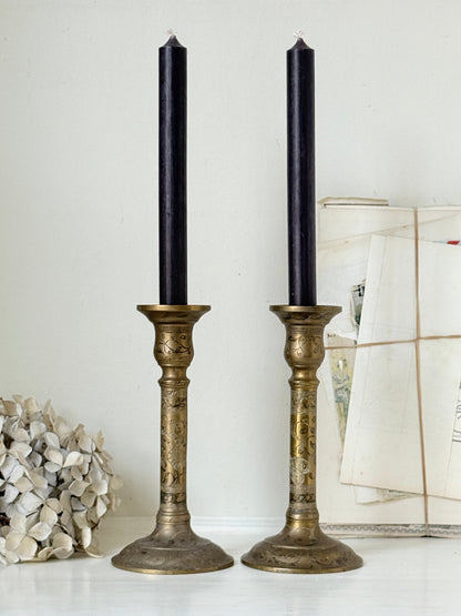 Pair of tall Indian engraved brass candlesticks in the Benares style with floral detailing and black taper candles on a white surface