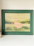 A framed folk art print featuring a pink goose standing in a green field with rolling hills and a small red house in the background, housed in a dark green wooden frame.