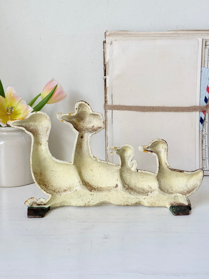 A vintage-style metal duck family sculpture in a distressed cream finish, displayed on a white tabletop next to a vase of tulips and a rustic framed print.