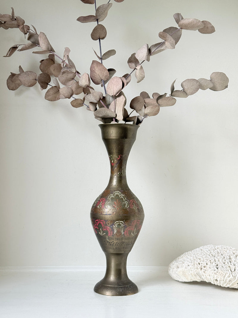 Vintage Indian engraved brass vase with enamel floral decoration and aged patina styled with dried stems on white surface