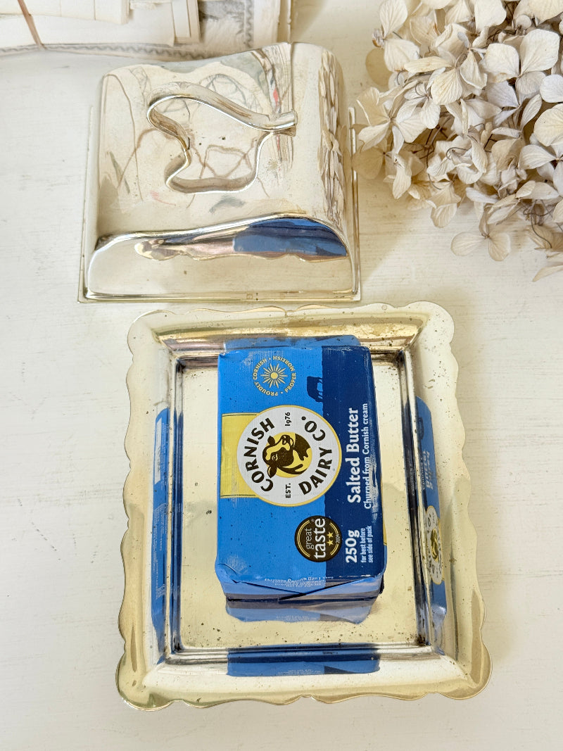 A vintage silver tray holding a blue and white package of Cornish Co. Dairy Salted Butter, with a second matching silver tray above it on a light surface next to dried flowers.