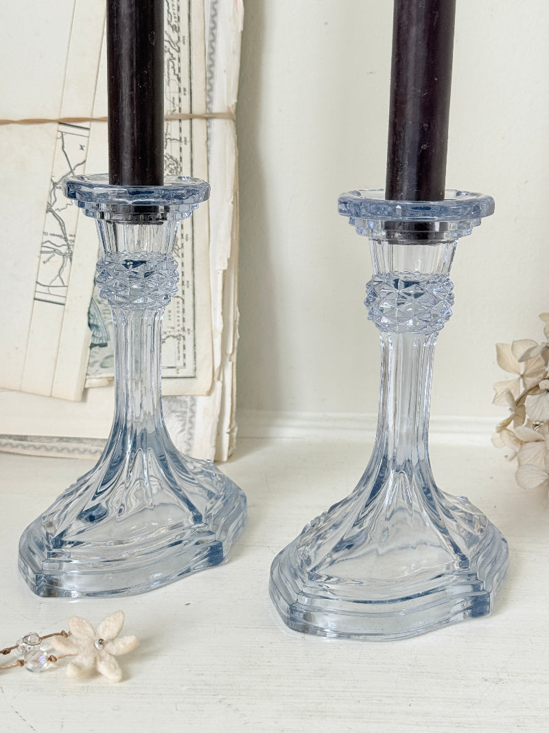Two clear glass candlestick holders with a light blue tint, shown on a white surface with dark candles and vintage papers in the background.