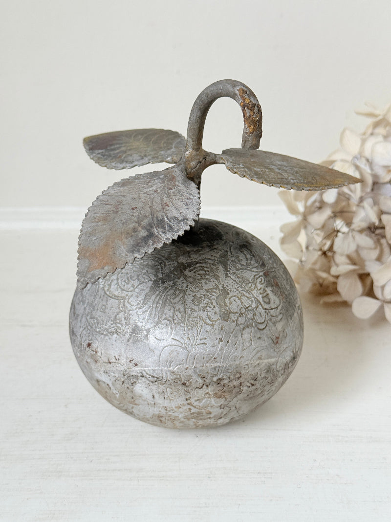 A decorative metal apple-shaped ornament with embossed floral patterns, a curved stem loop, and two leaf-like side extensions, shown on a light surface with a soft neutral background and dried flowers at the side.