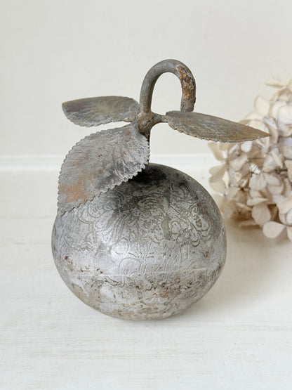 A decorative metal apple-shaped ornament with embossed floral patterns, a curved stem loop, and two leaf-like side extensions, shown on a light surface with a soft neutral background and dried flowers at the side.