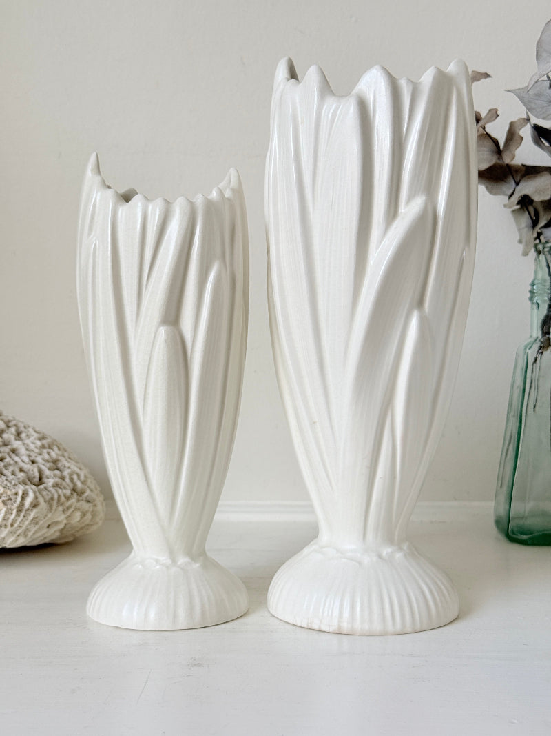 Two white ceramic tulip-shaped vases with ribbed stems and flared bases, displayed on a white surface against a light beige wall with decorative dried flowers and a green glass vase in the background.