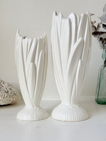 Two white ceramic tulip-shaped vases with ribbed stems and flared bases, displayed on a white surface against a light beige wall with decorative dried flowers and a green glass vase in the background.