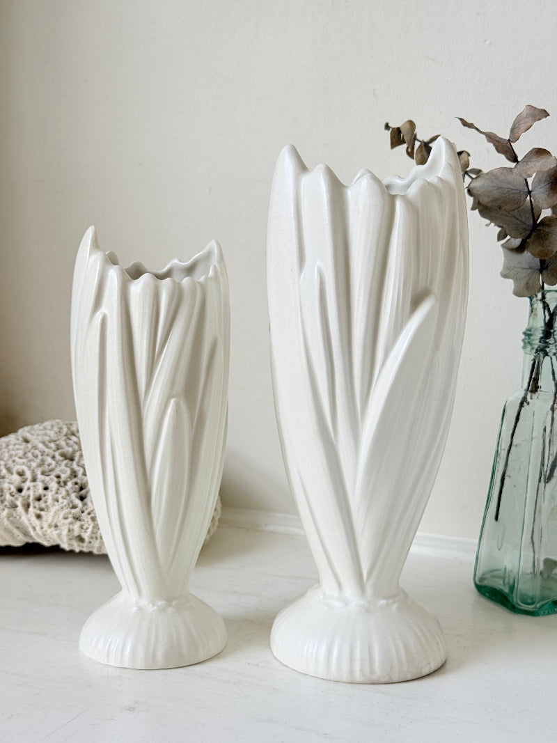 Two white ceramic tulip-shaped vases with fluted bases, displayed on a light surface against a neutral cream background, with a green glass vase and dried flowers as decorative props.