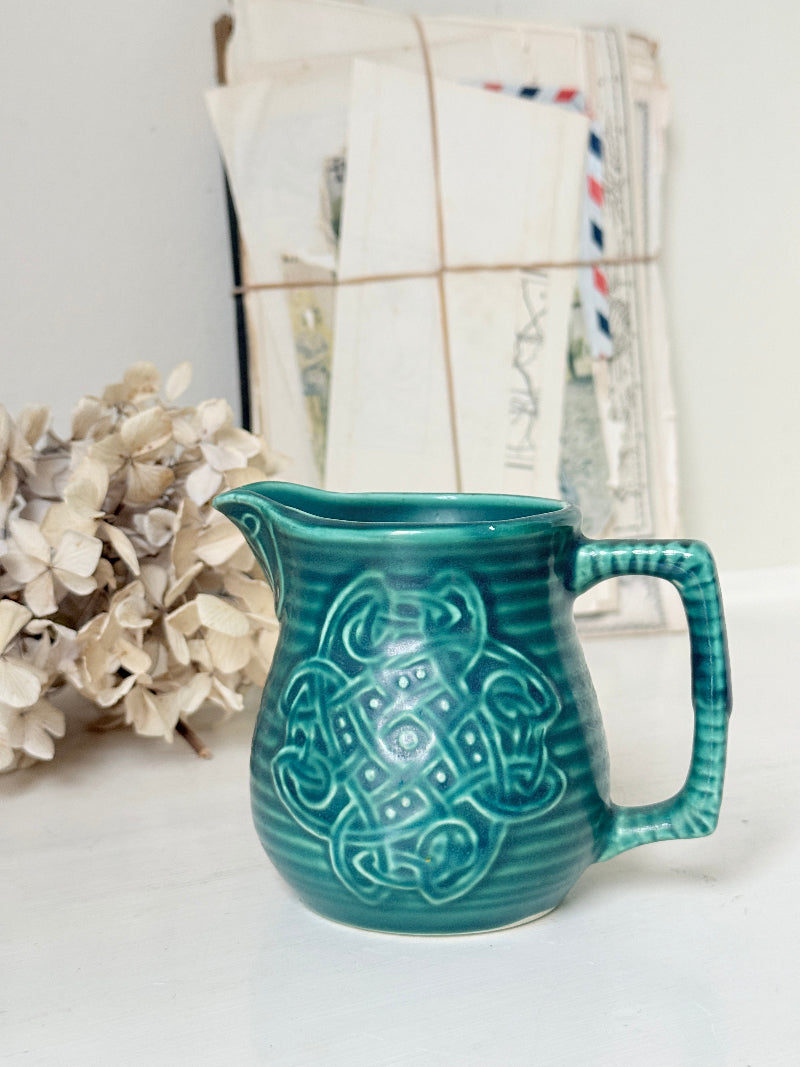 A teal ceramic pitcher with embossed Celtic knotwork patterns, styled on a white surface next to dried flowers and a bundle of vintage letters.