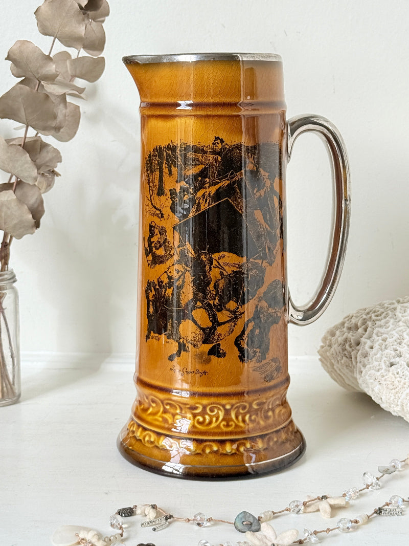 Antique Ridgways stoneware tall jug photographed on a white surface in amber treacle glaze with black transfer scenes and silver tone rim