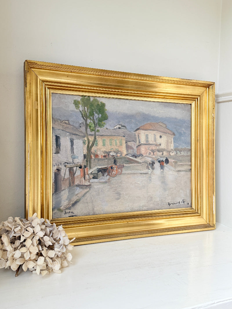 A framed impressionist landscape painting of a European town square, featuring a prominent gold frame, displayed on a white shelf next to dried white flowers.