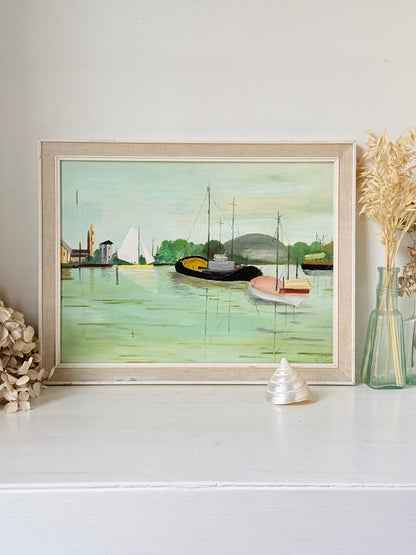 A framed art print depicting a serene harbor scene with boats on greenish water, styled on a white shelf with dried flowers and a small shell.