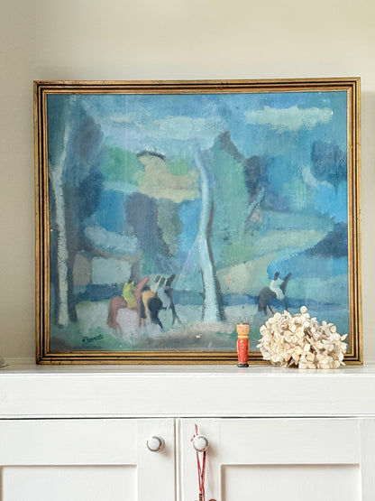 A framed landscape painting with blue, green, and gray tones, displayed on a white cabinet against a beige wall, next to a small red bottle and a white floral arrangement.