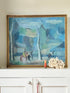 A framed landscape painting with blue, green, and gray tones, displayed on a white cabinet against a beige wall, next to a small red bottle and a white floral arrangement.